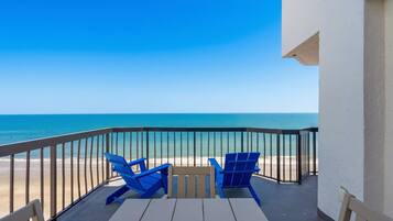 A balcony with a view of the beach and ocean, featuring two blue chairs and a table.
