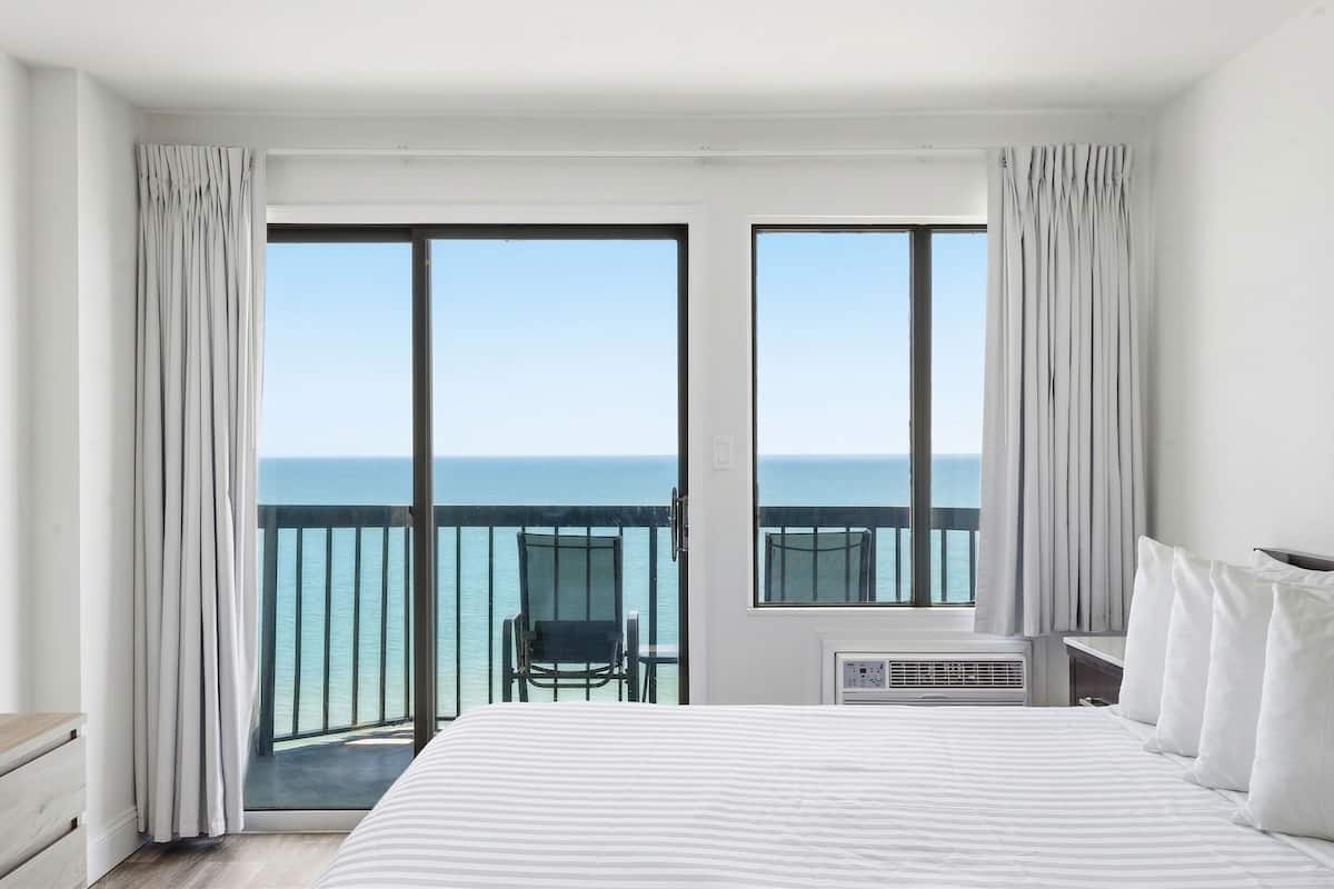 Habitación de hotel con cama, una silla, una mesita y vistas al océano a través de grandes ventanales.