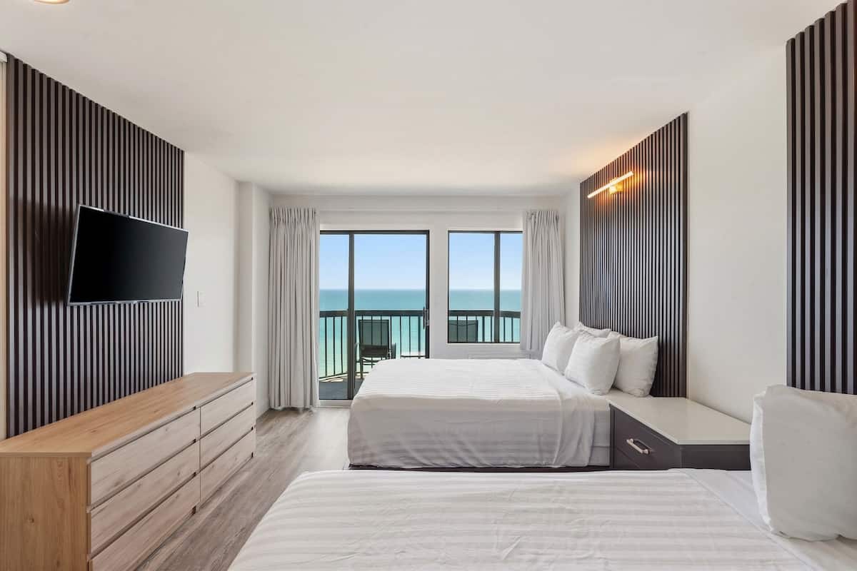 Habitación de hotel con dos camas, televisión, cómoda de madera y balcón con vista al mar.
