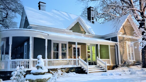A snow-covered house with a porch, a chimney, and a stone fountain. - The Gallery House Bed & Breakfast (Stephenville)