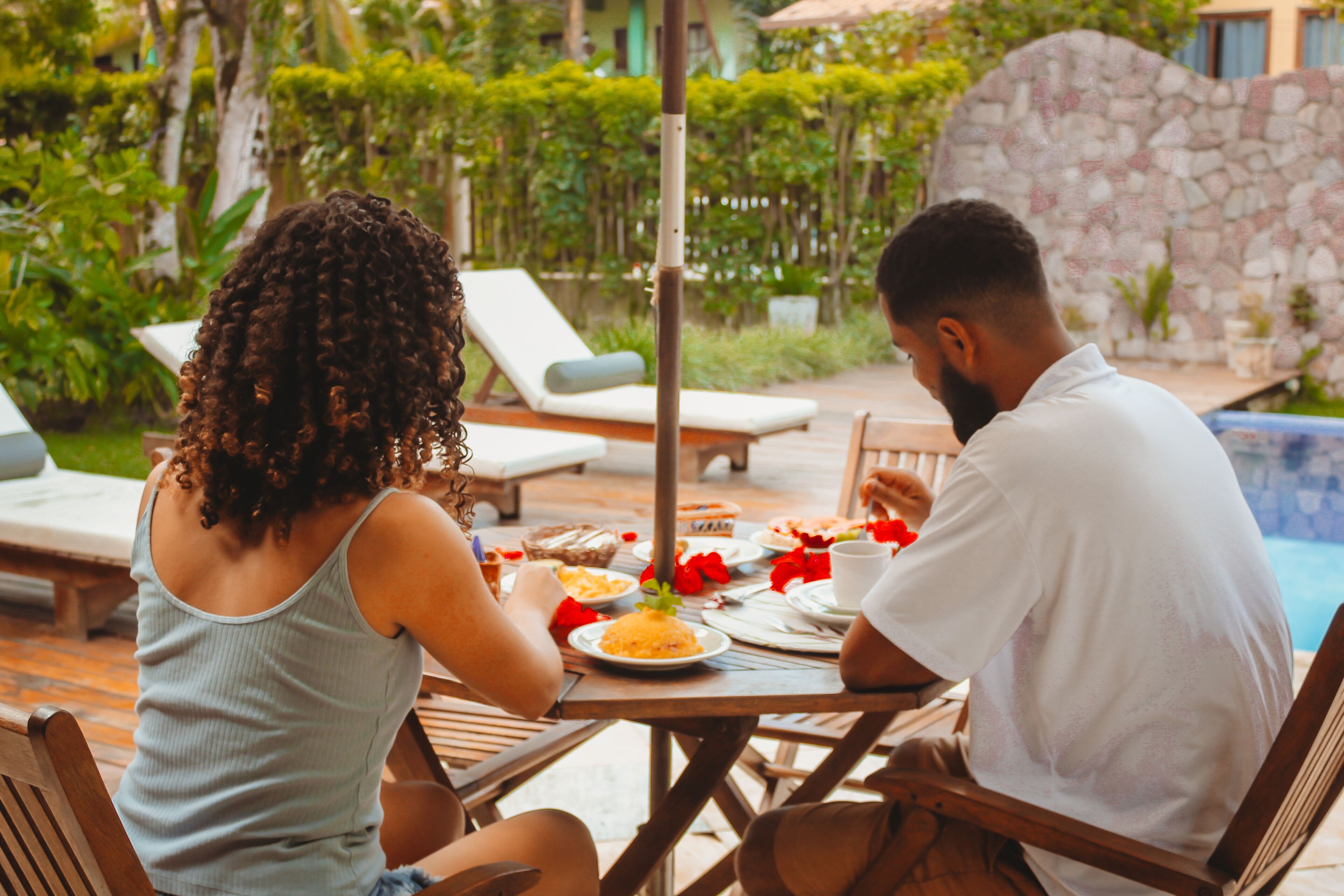 A couple dining at an outdoor table with a view of a swimming pool and lush greenery.