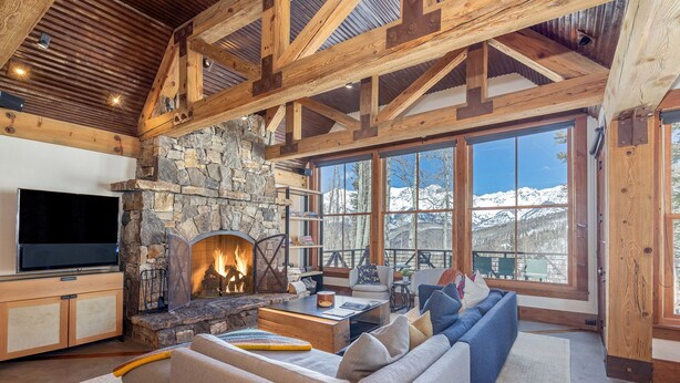 A cozy living room with a stone fireplace, large windows with a mountain view, and a flat-screen TV. - Luxury Home | Hot Tub, Decks + Sweeping Mountain views (Mountain Village)