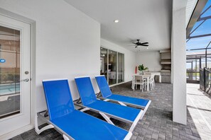 Un balcon avec des fauteuils de jardin bleus, un ventilateur de plafond et, au fond, un coin cuisine.