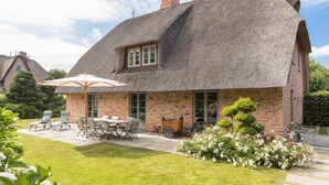 A thatched-roof house with a brick exterior, a patio with outdoor furniture, and a garden with flowers. - House Hansen Hus, Germany (Sylt)