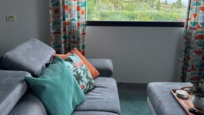 Una sala de estar moderna con sofás grises, cortinas coloridas con estampado y vistas a un paisaje verde a través de un ventanal grande.
