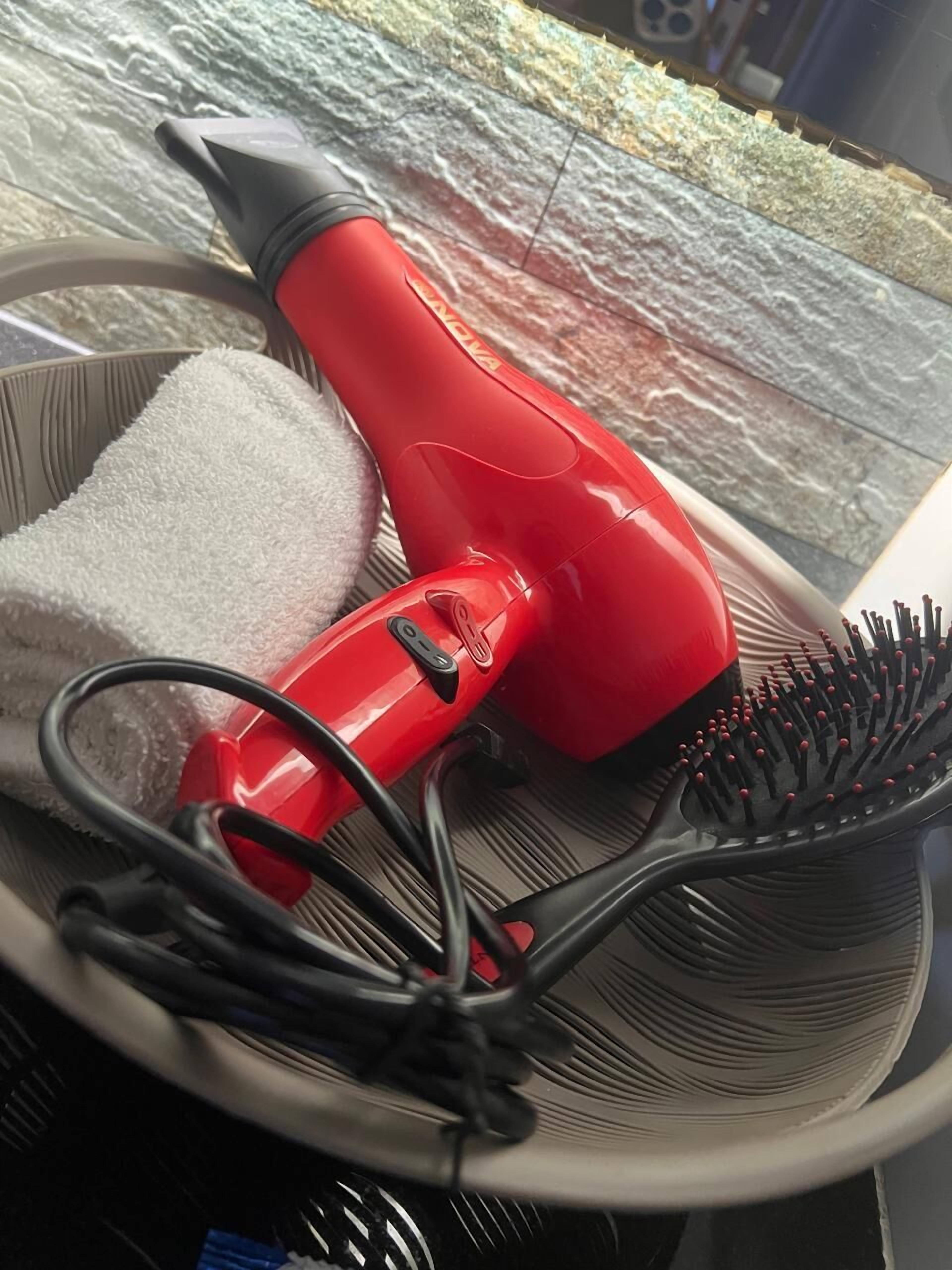 A red hairdryer, a white towel, and a black brush in a basket.
