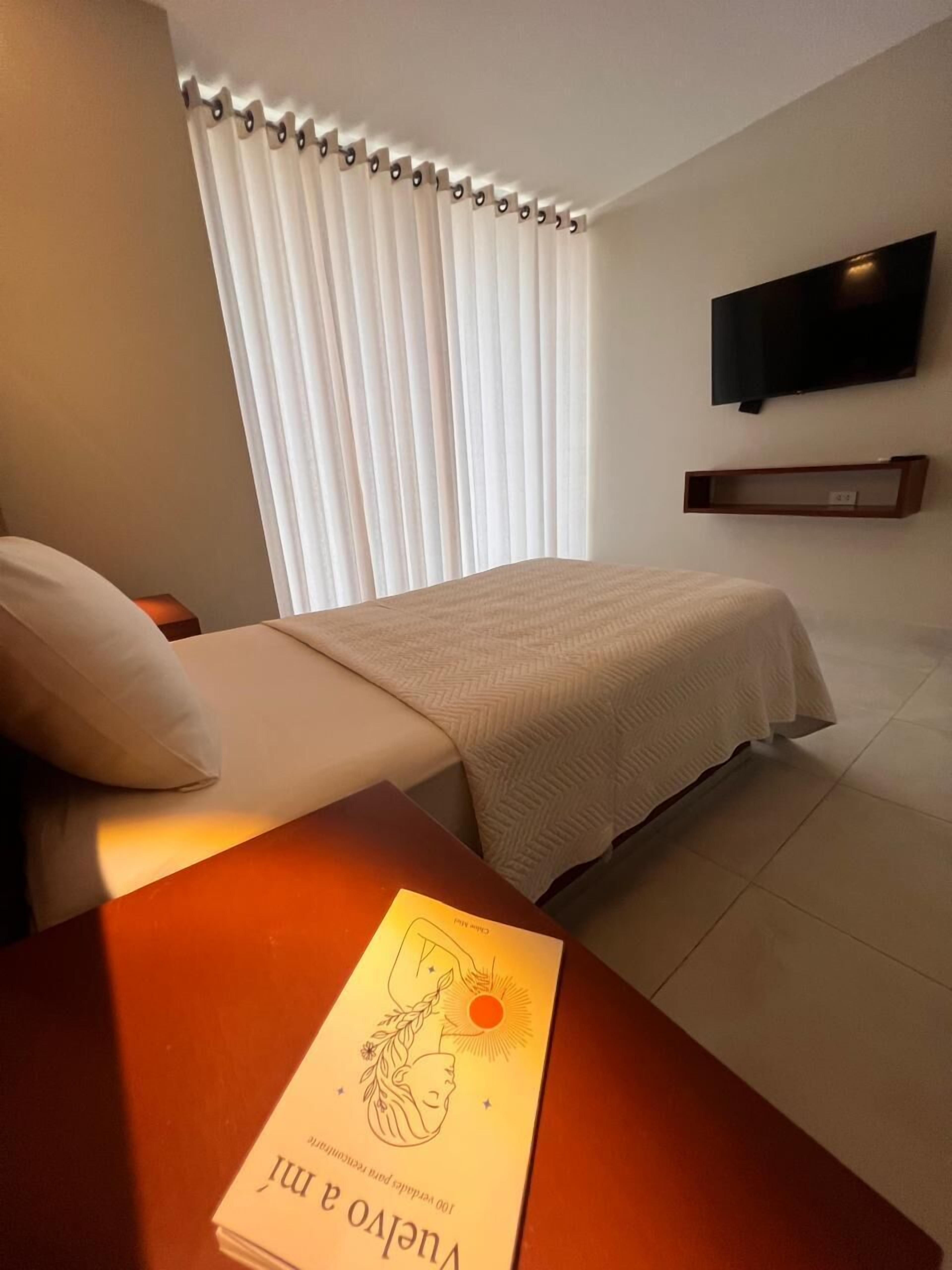 Habitación de hotel con cama, mesita de noche con un libro, televisor montado en la pared y ventana con cortinas.