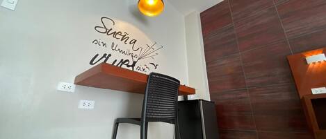 A modern kitchen with a black chair, a wooden shelf, a black refrigerator, and a wall-mounted light fixture.