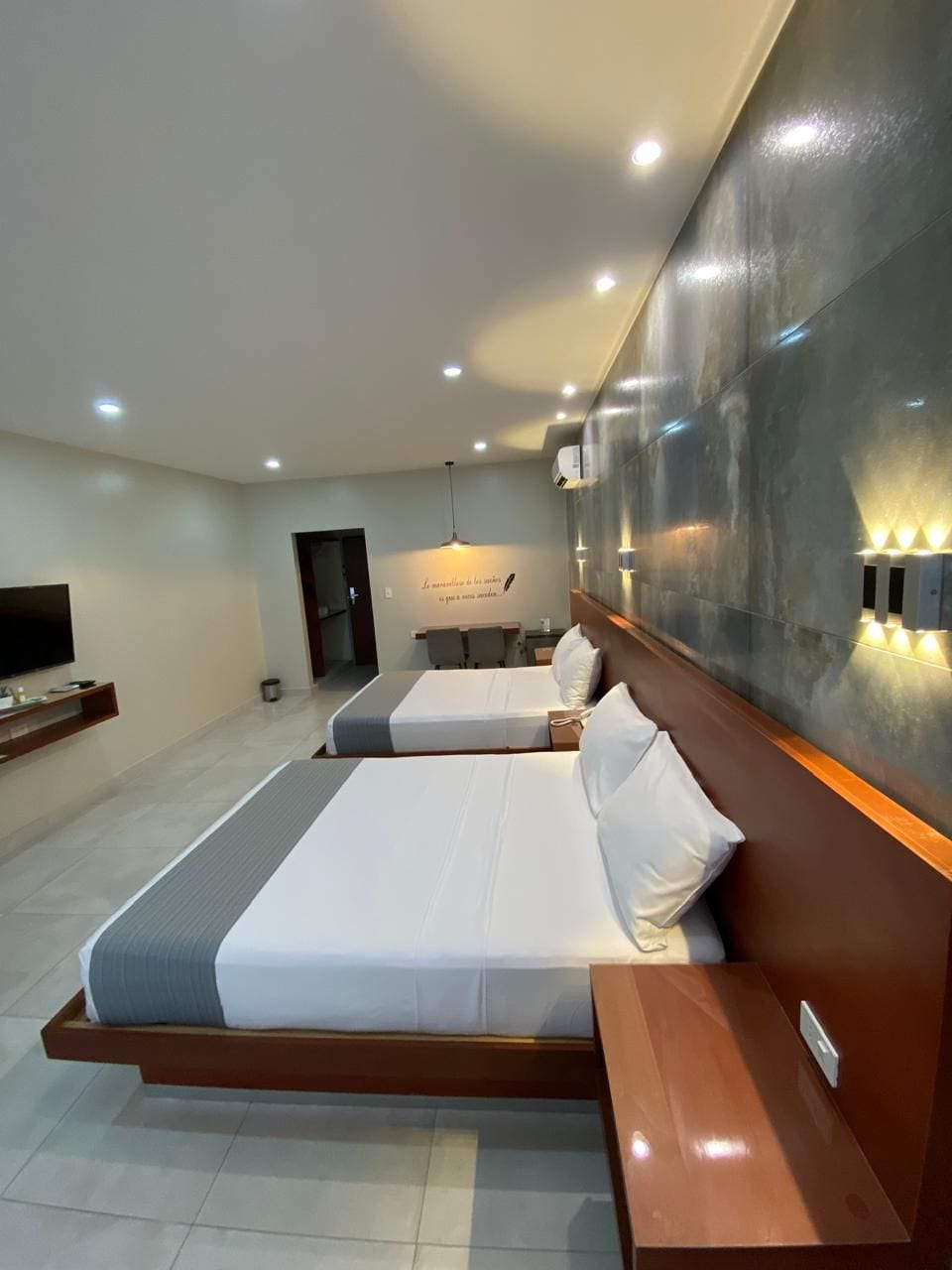 Een moderne hotelkamer met twee bedden, een televisie, wandlamp en een kitchenette op de achtergrond.