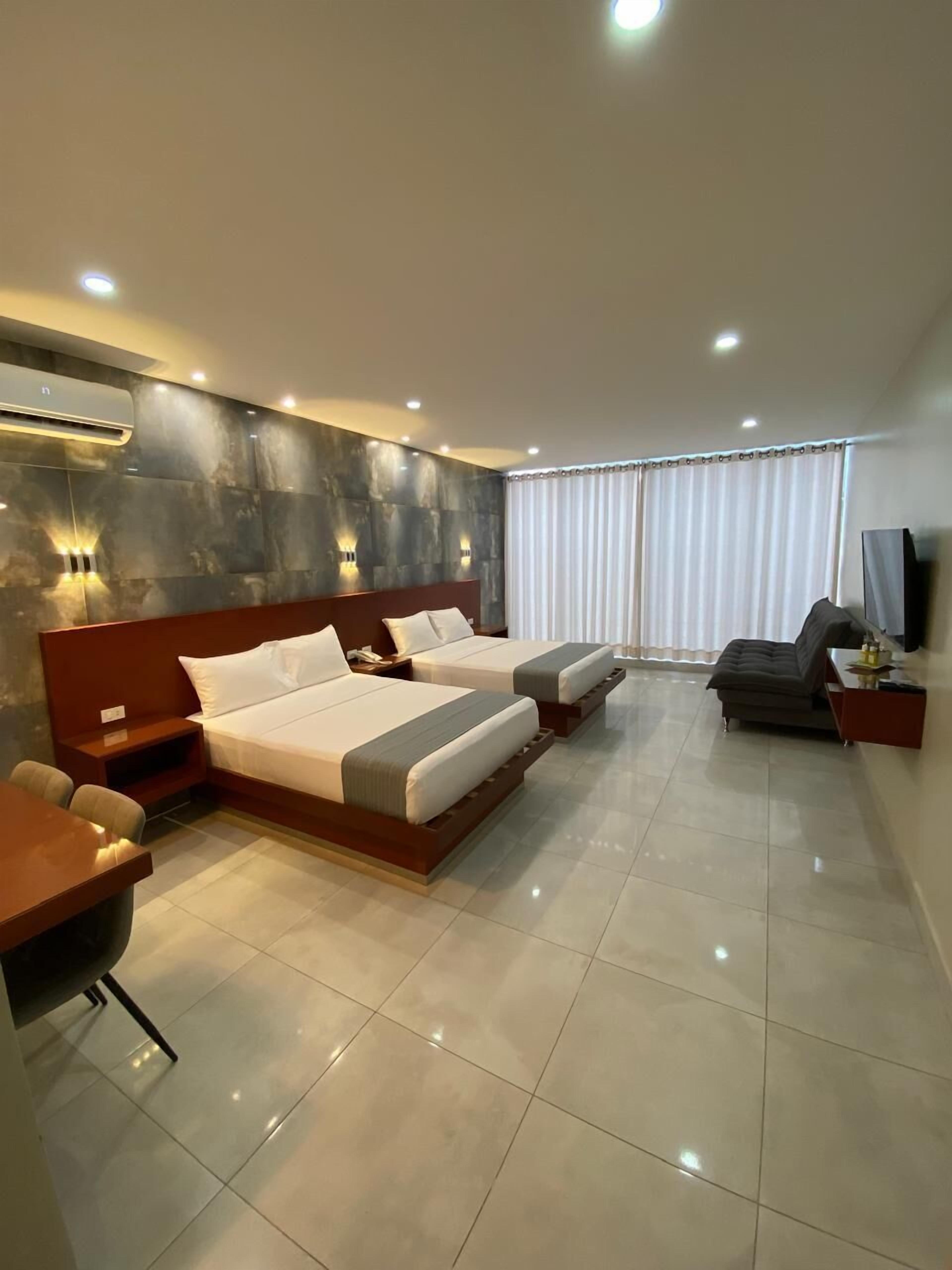 A hotel room with two beds, a desk, a chair, a TV, and a large window with curtains.