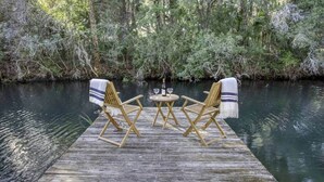 A wooden dock with two foldable chairs and a small round table, set up over a body of water surrounded by dense greenery. - River Rat's Rest (Homosassa)