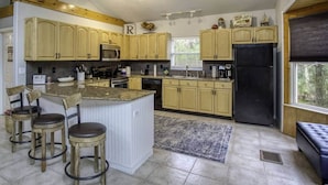 A kitchen with light wooden cabinets, a central island with stools, a black refrigerator, and a window with blinds. - River Rat's Rest (Homosassa)