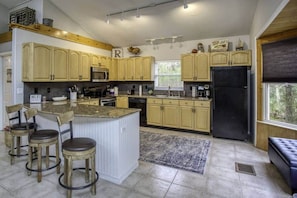 A kitchen with light wooden cabinets, a central island with stools, a black refrigerator, and a window with blinds. - River Rat's Rest (Homosassa)