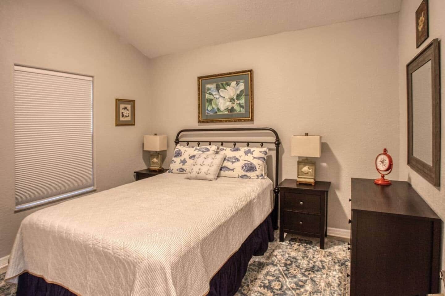 A bedroom with a bed, two bedside tables, a nightstand, a window with blinds, a framed picture on the wall, and a clock on the nightstand.