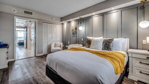 A bedroom with a large bed, a bedside table, a chair, and a wardrobe. There is a door leading to another room. - Spectacular Beach Condo Double Suite for 2 couples (Myrtle Beach)