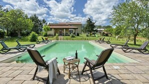 Piscina retangular com espreguiçadeiras e uma mesa, cercada por um jardim ajardinado e um edifício ao fundo.
