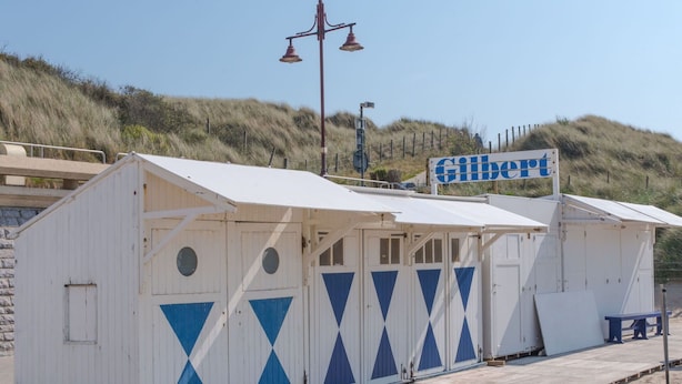 Ein Strandhäuschen namens 'Gilbert' mit weißer Fassade und blauen geometrischen Mustern, gelegen an einem Sandstrand mit einer Holzterrasse und einem Laternenpfahl.