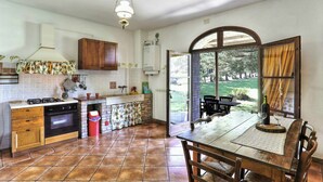 A kitchen with a gas stove, oven, and wooden cabinets. There is a dining table with chairs and a view of the outdoors through a large window. - Lovely apartment in villa for 5 guests with pool, WIFI, TV, terrace and panoramic view (Pomarance)