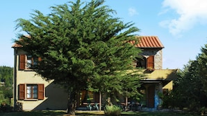 A two-story house with a stone facade, wooden shutters, and a large tree in the front yard. - Beautiful apartment in villa for 5 people with WIFI, pool, TV and veranda (Pomarance)