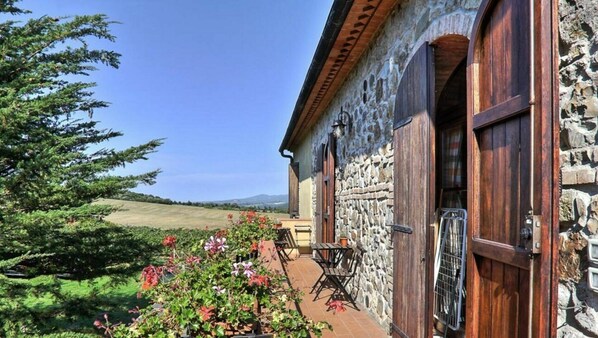 A stone building with wooden shutters, a patio with a table and chairs, and a view of a green landscape.