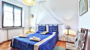A bedroom with a large bed, a window with curtains, a bedside table with a lamp, and a chair with a floral cushion. - Beautiful apartment in villa for 5 people with WIFI, pool, TV and veranda (Pomarance)
