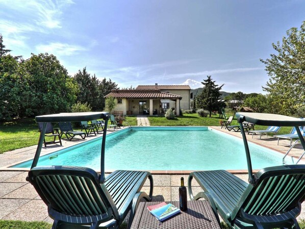 A rectangular swimming pool with lounge chairs on either side, a small building in the background, and a well-maintained lawn.
