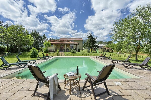A pool area with lounge chairs and a table, surrounded by a garden and a building with a tiled roof.