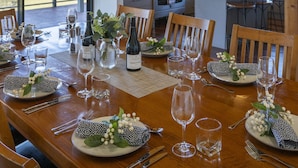 A dining area with a wooden table set for a meal, a kitchen in the background, and large windows offering a view of the outdoors. - Old Coach Road Estate (Hindmarsh Valley)