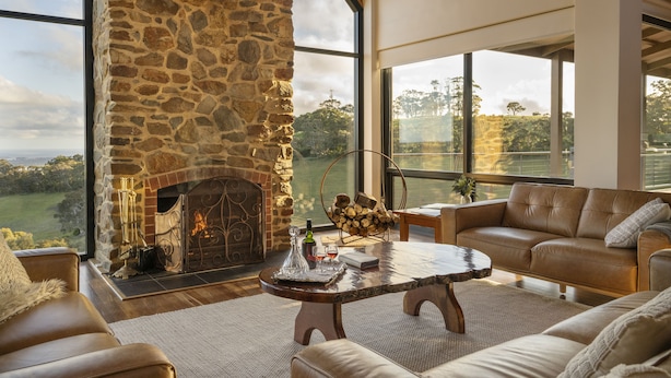 A spacious living room with a stone fireplace, leather sofas, and large windows offering a view of the countryside. - Old Coach Road Estate (Hindmarsh Valley)