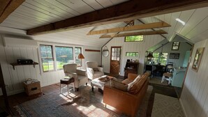 Ein großzügiger Wohnbereich mit hoher Decke, Holzbalken und großen Fenstern. Man sieht ein Sofa, Sessel, einen Tisch und eine Küchenzeile.