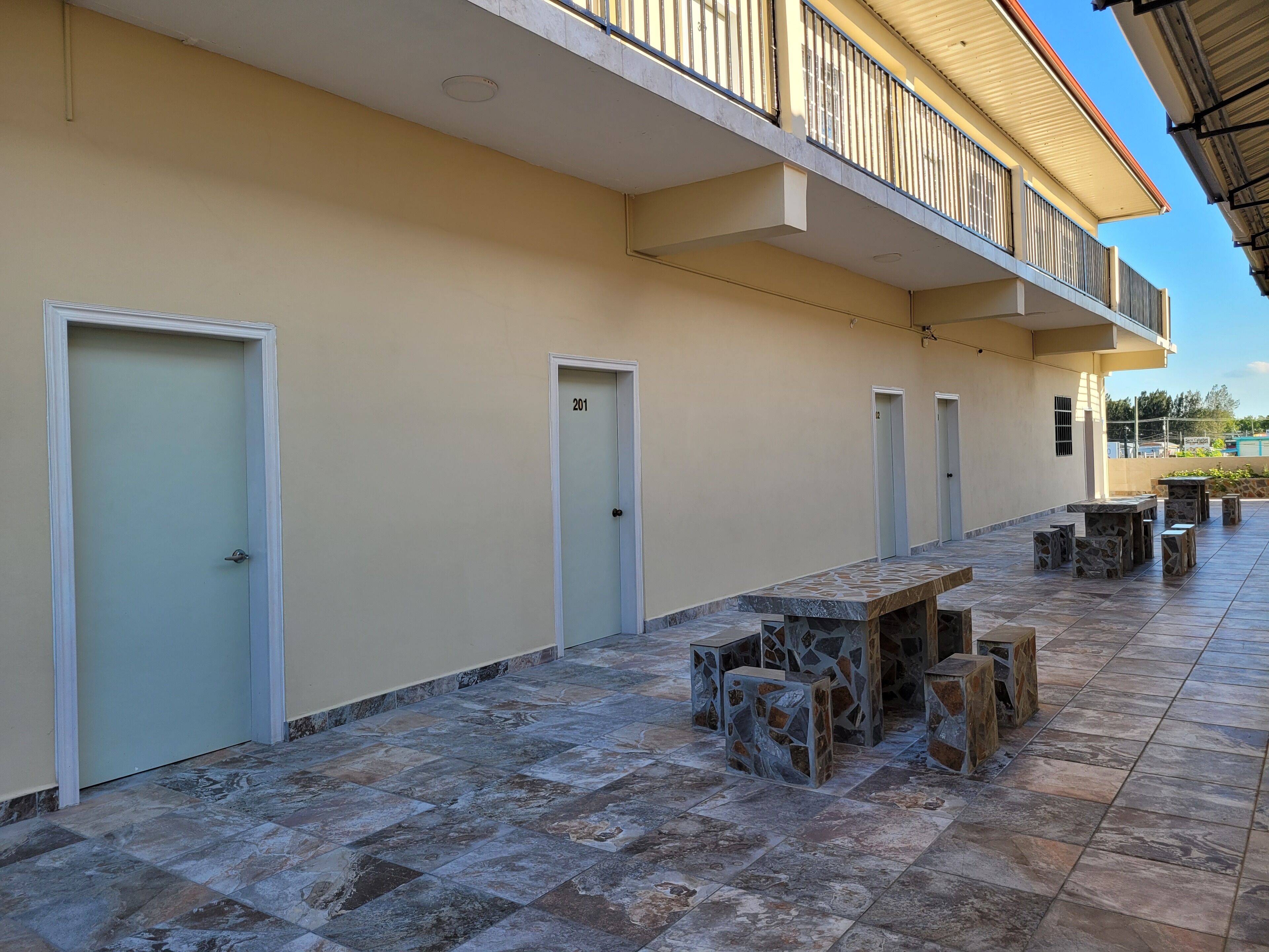 Un bâtiment doté d’un toit plat, d’un balcon avec des rampes en métal et d’une terrasse carrelée avec des tables et des chaises en pierre.