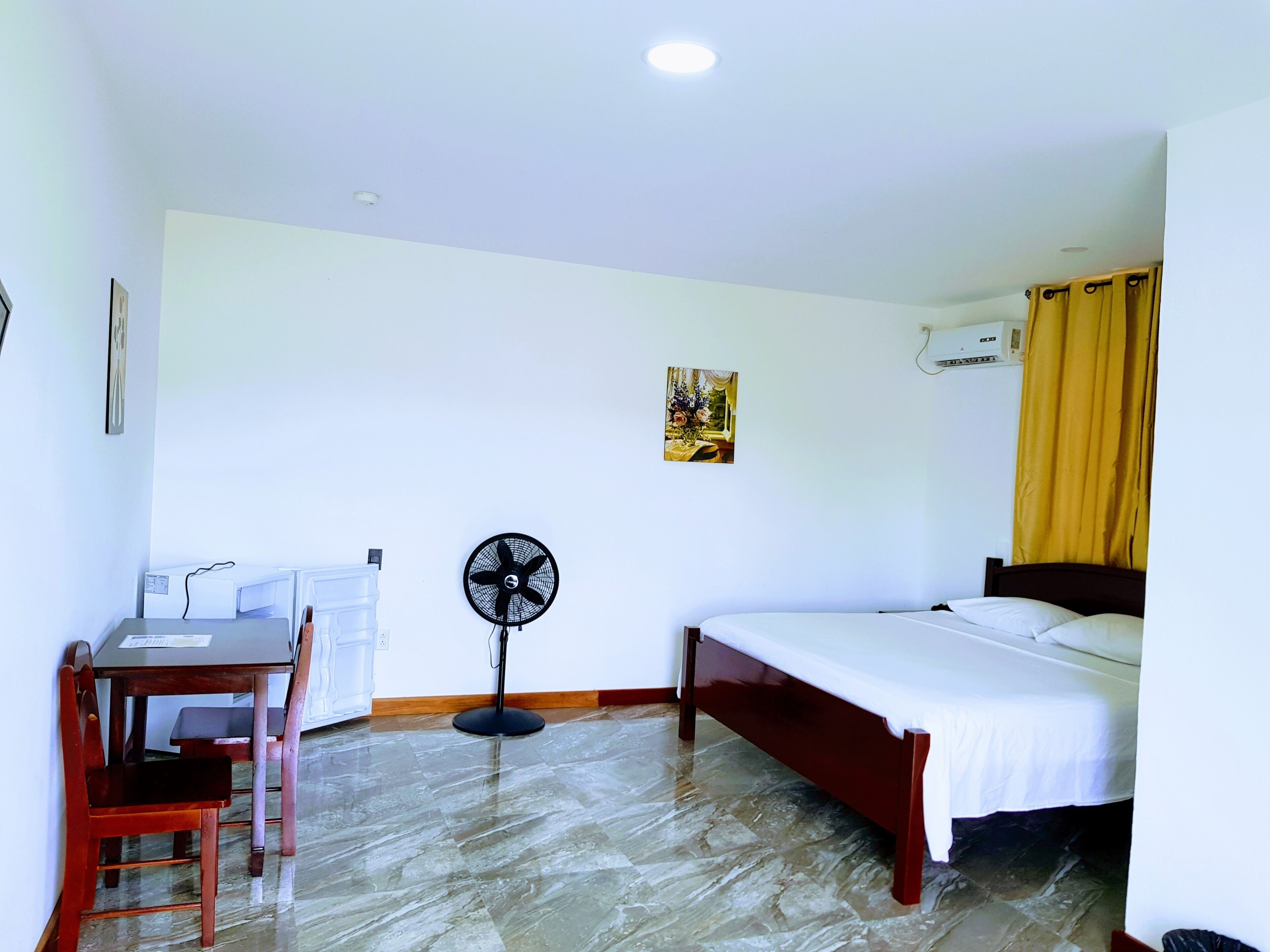 A room with a bed, a desk with a chair, a standing fan, and an air conditioning unit.