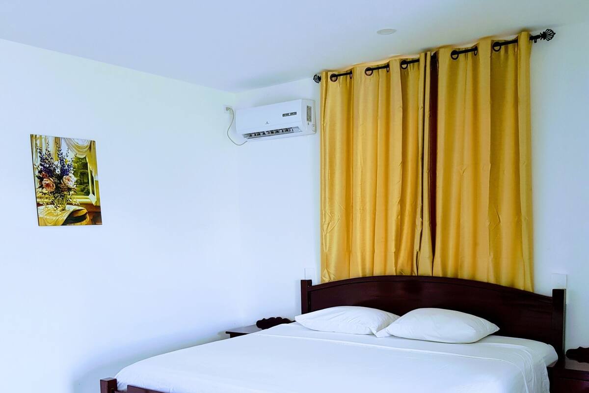 A bedroom with a bed, a nightstand, a wall-mounted air conditioner, and a window with curtains.