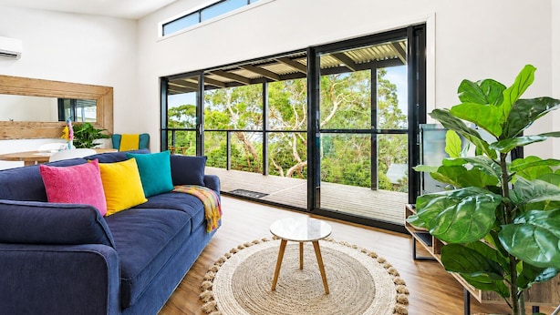 A modern living room with a blue sofa, colorful cushions, a round coffee table, and a large sliding glass door leading to a balcony with a view of trees. - Top of the Bay by Ready Set Stay (Arthurs Seat)