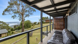 A covered outdoor area with a wooden deck, a black leather sofa, and a view of the sea and hills. - Top of the Bay by Ready Set Stay (Arthurs Seat)
