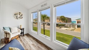 A room with a sofa, a small table, and a wall-mounted decoration. There are three large windows offering a view of the beach and the ocean.