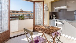 A balcony with a wooden table and chairs, a kitchen area with modern appliances, and a view of a building across the street. - Le Grand Large by Interhome (Le Lavandou)