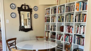 A dining area with a round table, four wooden chairs, a patterned rug, a bookshelf filled with books, a mirror, and decorative plates on the wall.