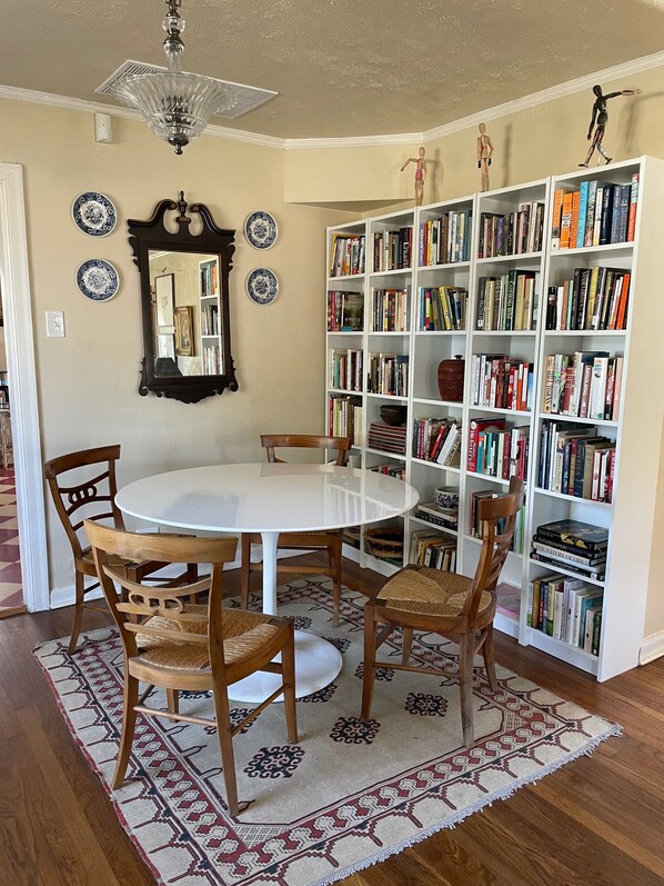 A dining area with a round table, four wooden chairs, a patterned rug, a bookshelf filled with books, a mirror, and decorative plates on the wall.
