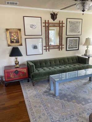 A living room with a green sofa, a glass coffee table, a red chest of drawers, and various framed artworks on the wall.