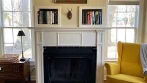 A living room with a fireplace, bookshelves, a yellow armchair, a bench, and a patterned rug.
