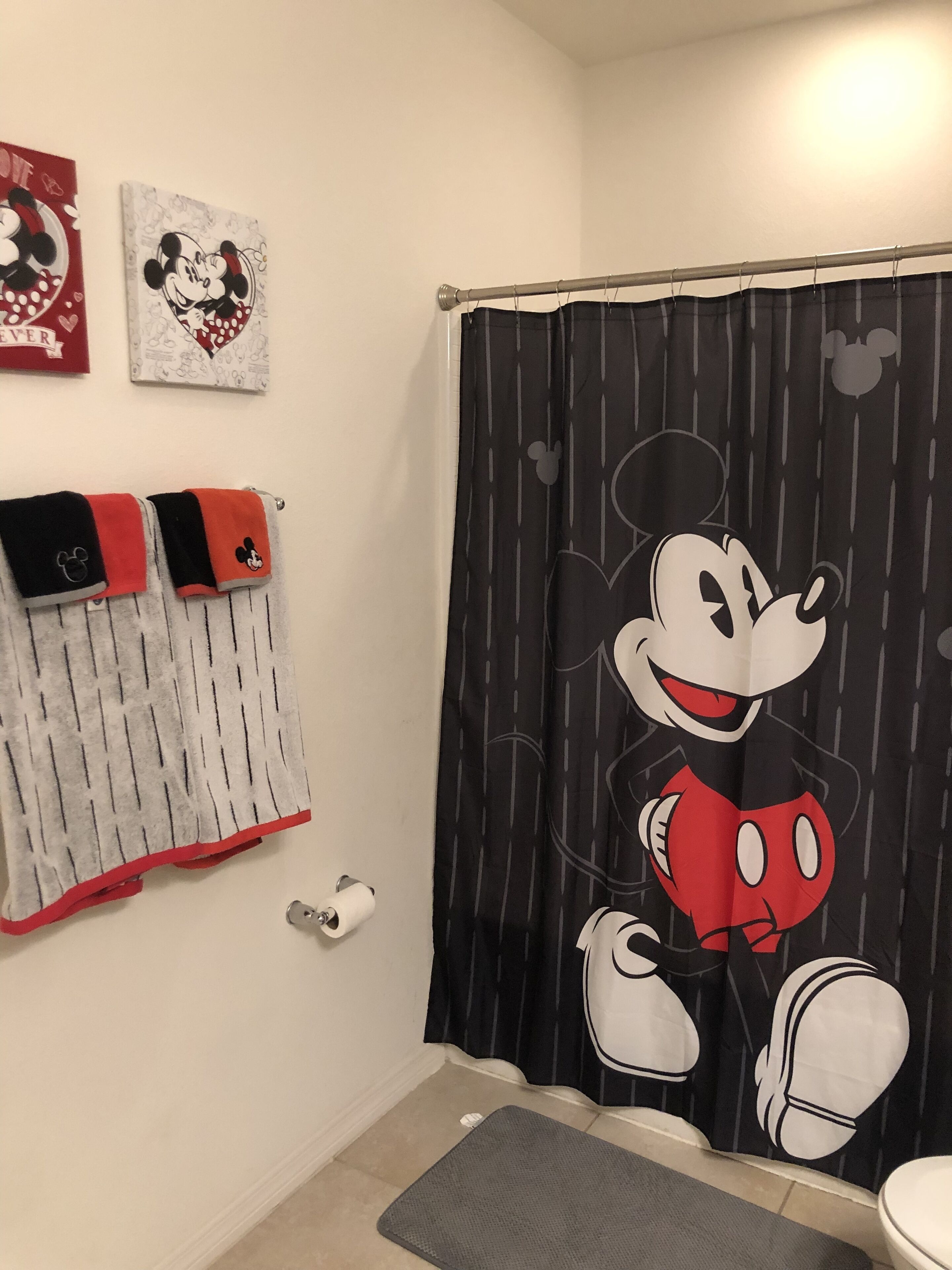 Ein Badezimmer mit Mickey Mouse Duschvorhang, einem Handtuchhalter mit Mickey-Motivhandtüchern und einem kleinen Teppich auf dem Boden.