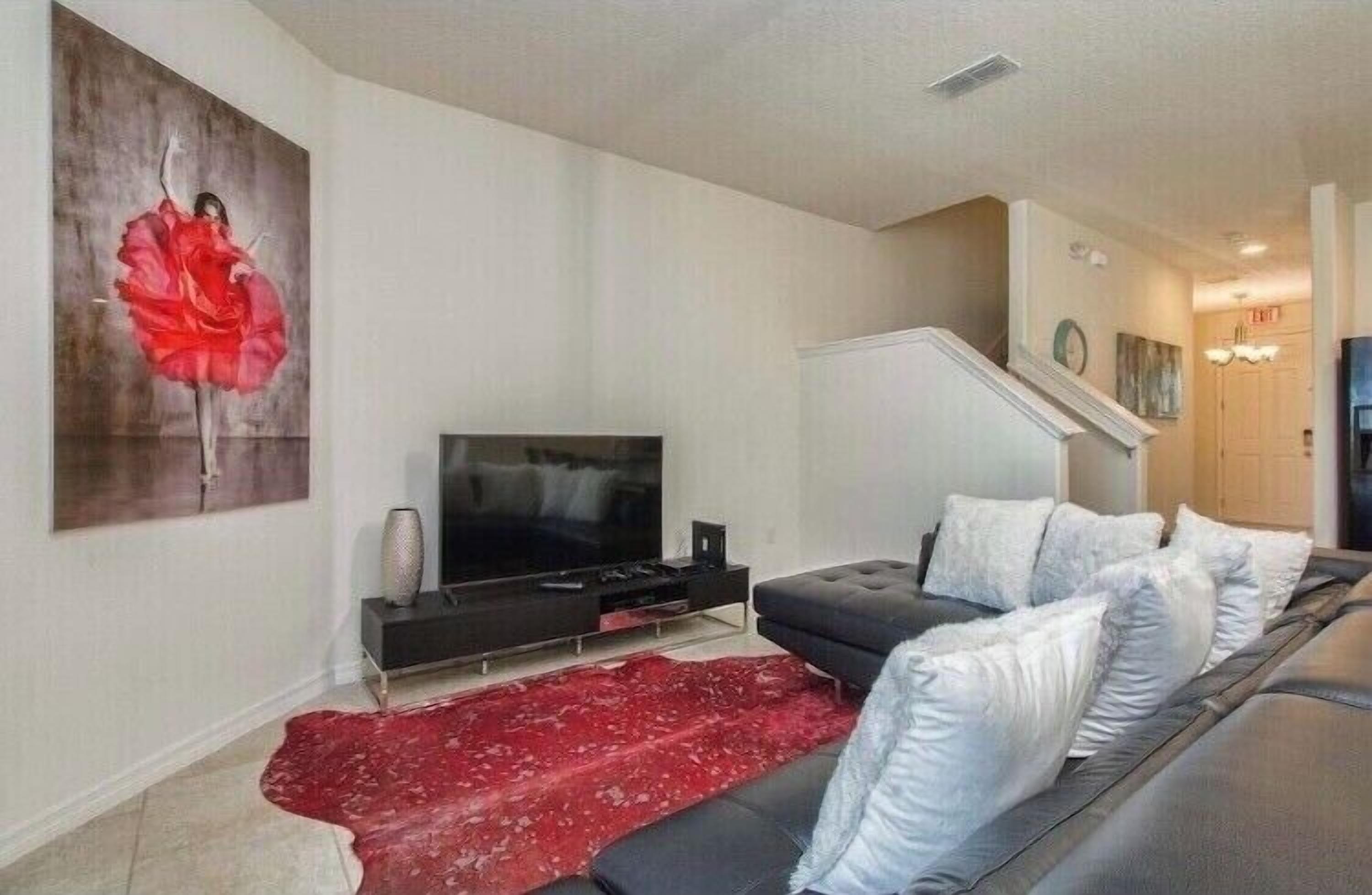 A modern living room with a flat-screen TV, a black sofa with white cushions, a red rug, and a large abstract painting on the wall.