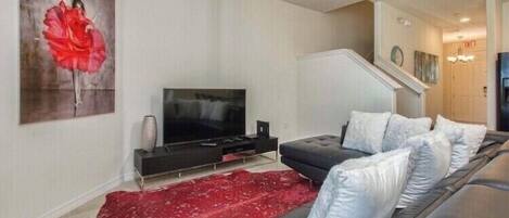 A modern living room with a flat-screen TV, a black sofa with white cushions, a red rug, and a large abstract painting on the wall.