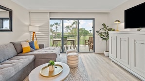 Una sala moderna con un sofá seccional gris, una mesa de centro con una bandeja y una planta, un área de comedor con sillas y una mesa, un armario blanco y una puerta corrediza de vidrio que da a un balcón con mobiliario exterior.