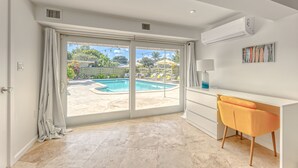 A spacious room with a large sliding glass door leading to a pool area, a white desk with a lamp, and a modern chair. - Newly Renovated Beach House w Heated Pool, 4 BR, 3 BA (Fort Lauderdale)