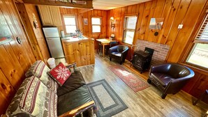 A cozy cabin interior with a wooden floor, a sofa with patterned cushions, a leather armchair, a dining table, and a fireplace. - Cozy Cabin with Lake Views - Rustic Getaway (Indian Lake)