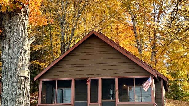 A small cabin with a gabled roof, large windows, and an American flag on the front. - Cozy Cabin with Lake Views - Rustic Getaway (Indian Lake)