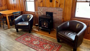 A cozy room with wooden walls, a fireplace, a dining table, two leather chairs, and a red patterned rug. - Cozy Cabin with Lake Views - Rustic Getaway (Indian Lake)
