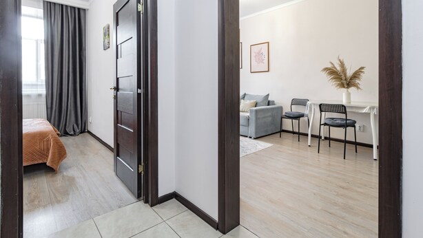 Uma sala de estar moderna com sofá, mesa de jantar e cadeiras. Há uma porta que dá para outro quarto com uma cama.